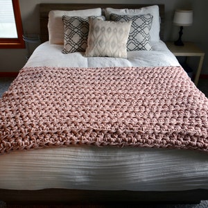 May include: A blush pink crocheted throw blanket on a white bed with patterned throw pillows.
