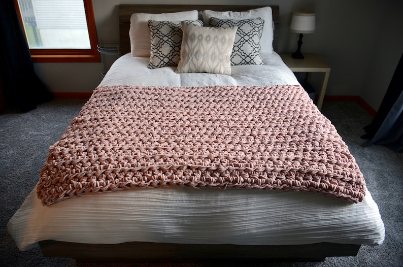May include: A blush pink crocheted throw blanket on a white bed with patterned throw pillows.