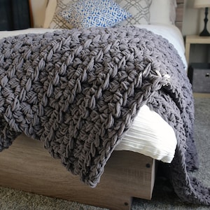May include: A gray chunky knit throw blanket draped over a bed with white bedding. The blanket has a textured, woven pattern.