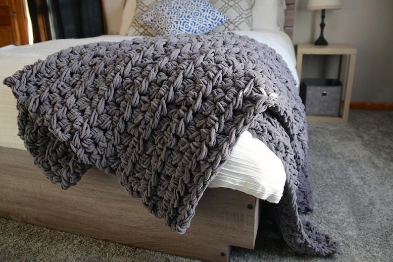 May include: A gray chunky knit throw blanket draped over a bed with white bedding. The blanket has a textured, woven pattern.