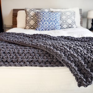 May include: A gray crocheted throw blanket draped over a white bed with patterned pillows. The blanket is made with thick yarn and has a textured, chunky look.
