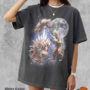 Puede incluir: Una camiseta de manga corta color pimienta con un estampado gráfico de dos monstruos reptiles con espinas en la espalda, uno de pie y otro en primer plano, sobre un fondo espacial estrellado con una gran luna llena y rayos de luz blancos.