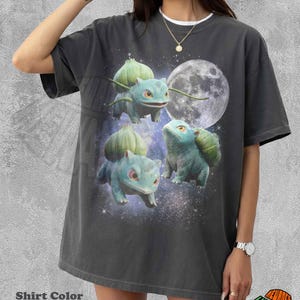 Puede incluir: Una camiseta de manga corta color pimienta con un estampado gráfico de tres criaturas Bulbasaur verdes sobre un fondo espacial oscuro con una luna grande. El diseño incluye un motivo de cielo estrellado que rodea a las criaturas en el frente.