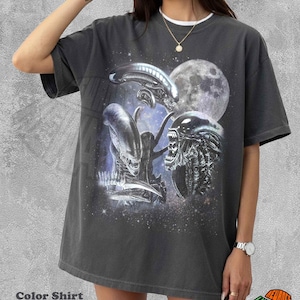 Puede incluir: Una camiseta gris carbón de manga corta con un estampado gráfico de tres criaturas extraterrestres y una luna grande sobre un fondo espacial estrellado. La camiseta tiene cuello redondo y un corte relajado, con el texto Color Shirt Pepper.
