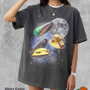 May include: A pepper-colored short-sleeve t-shirt featuring a graphic print of three open seashells with iridescent yellow and green interiors set against a dark space background with a large moon and scattered stars. The shirt has a crew neckline.