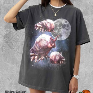May include: Pepper-colored t-shirt featuring a graphic of three alien-like creatures with a full moon and starry background. The shirt has a white collar and the text "Shirt Color Pepper".