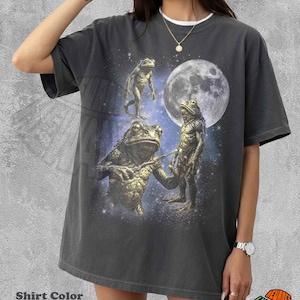May include: Pepper-colored t-shirt featuring a surreal graphic of anthropomorphic frogs under a full moon. The design includes three frog figures, a starry background, and the text "REWARDS". The shirt has a relaxed fit.