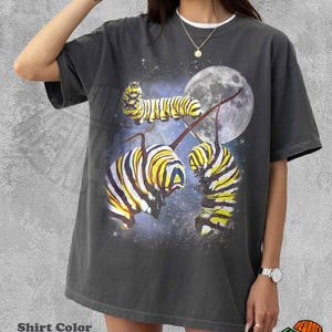 May include: Pepper-colored t-shirt featuring a graphic of caterpillars against a starry night sky with a full moon. The caterpillars are yellow and black. The shirt has a white collar. The text "Shirt Color Pepper" is at the bottom.
