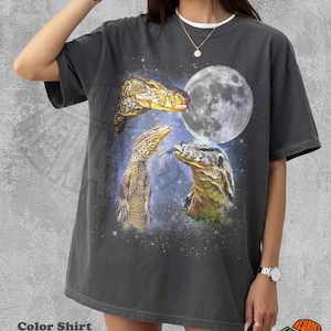 May include: A charcoal gray short-sleeve t-shirt featuring a graphic print of three yellow and black monitor lizards against a starry night sky background with a large full moon. The shirt has a crew neckline and a relaxed, loose-fitting silhouette.