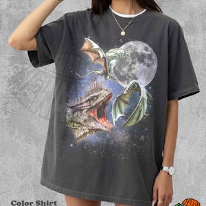 May include: Dark grey t-shirt with a fantasy dragon design, a full moon, and a starry background. The shirt has a white crew neck collar and the text "Color Shirt Pepper". The shirt is a relaxed fit.