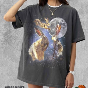 May include: A pepper-colored short-sleeve t-shirt featuring a graphic print of three brown rabbits set against a starry night sky with a large full moon. The design includes one rabbit leaping, one sitting, and one large rabbit head in the foreground.