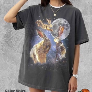 May include: A charcoal gray short-sleeve t-shirt featuring a graphic print of three brown rabbits against a backdrop of a full moon and a starry night sky. The shirt has a crew neckline and a relaxed fit, with the text Color Shirt Pepper visible.