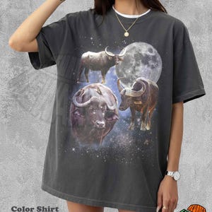 May include: A pepper-colored short-sleeve t-shirt featuring a graphic print of three buffaloes set against a starry night sky and a large full moon. The design includes a celestial theme with scattered stars and dark blue and black tones.