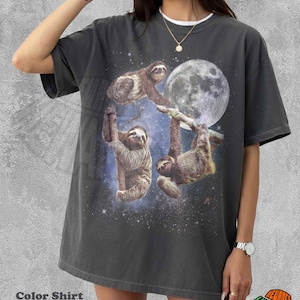 Puede incluir: Una camiseta de manga corta color gris carbón con un estampado gráfico de tres perezosos colgados de ramas sobre un fondo de luna llena y una galaxia estrellada. La prenda tiene cuello redondo y un corte holgado y relajado.
