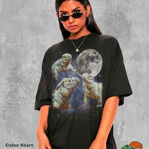 Puede incluir: Una camiseta negra de corte oversize con un estampado gráfico de tres iguanas sobre un fondo de cielo nocturno estrellado con una luna llena grande y detallada. El diseño incluye un tema celestial con estrellas dispersas alrededor de los reptiles.