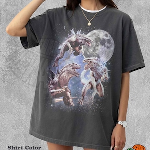 Può includere: Una t-shirt a maniche corte color pepe con una stampa grafica di tre creature simili a dinosauri su uno sfondo spaziale stellato con una grande luna. La maglietta presenta un girocollo e una vestibilità ampia e rilassata.