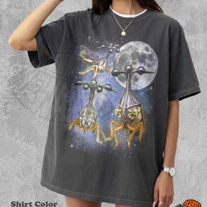 May include: A pepper-colored t-shirt featuring a graphic print of three alien-like insect creatures against a backdrop of a full moon and a starry purple nebula. The shirt has a crew neckline and short sleeves, showcasing a detailed space-themed design.