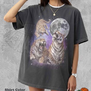 May include: A pepper-coloured short-sleeve t-shirt featuring a graphic print of two leopards and a tiger against a purple galaxy background with a large full moon. The design includes a leaping leopard, a roaring leopard, and a tiger looking upward.