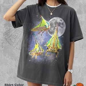 May include: A pepper-colored t-shirt featuring a graphic print of three green and yellow thorn bugs against a dark, starry space background with a full moon. The shirt has a crew neckline and short sleeves, showcasing a detailed insect illustration.
