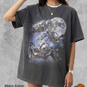 Puede incluir: Una camiseta de manga corta color pimienta con un estampado gráfico de tres criaturas metálicas similares a insectos con ojos verdes brillantes sobre un fondo de una luna grande y una galaxia estrellada en azul oscuro.