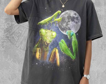 May include: A charcoal grey t-shirt featuring a graphic print of three bright green praying mantises against a dark, starry space background with a large, detailed full moon. The shirt has a crew neckline and short sleeves, showcasing a celestial theme.
