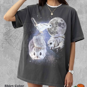 以下が含まれることがあります： ペッパーカラーの半袖Tシャツ。大きな月と星空の宇宙空間に、3匹のモモンガが描かれたグラフィックプリントが特徴です。滑空する姿、座っている姿、正面を向いている姿の3匹が、暗い宇宙の背景に配置されています。