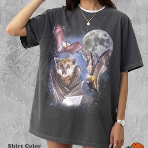 May include: A pepper-colored short-sleeve t-shirt featuring a graphic print of three bats in a starry night sky with a large full moon. The central bat is shown in a front-facing portrait style, while the other two bats are depicted in flight.