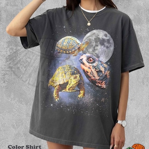 Op de afbeelding: Een antracietgrijs T-shirt met korte mouwen en een grafische print van twee schildpadden en een grote schildpaddenkop tegen een sterrenhemel met een volle maan. Het T-shirt heeft een ronde hals en een losse, ontspannen pasvorm.