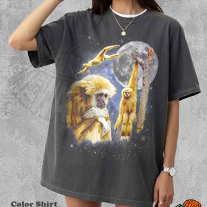 May include: A charcoal grey short-sleeve t-shirt featuring a graphic print of three yellow gibbons in a starry night sky with a large full moon. The design includes one gibbon leaping, one hanging from a tree branch, and one sitting in profile.