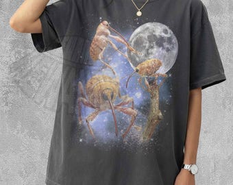 Puede incluir: Una camiseta de manga corta color pimienta con un estampado gráfico de tres gorgojos sobre un fondo de luna llena y cielo estrellado. La prenda tiene cuello redondo y un corte holgado, mostrando una ilustración detallada con temática espacial.