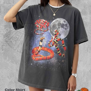 May include: A charcoal gray short-sleeve t-shirt featuring a graphic print of three snakes in red, black, and yellow patterns against a cosmic space background with a large full moon. The shirt has a crew neckline and a relaxed, boxy fit.