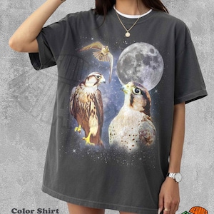 Może przedstawiać: Ciemnoszara koszulka z grafiką przedstawiającą trzy sokoły, pełnię księżyca i gwiezdne tło. Koszulka ma biały kołnierzyk i napis "Color Shirt Pepper" na dole.