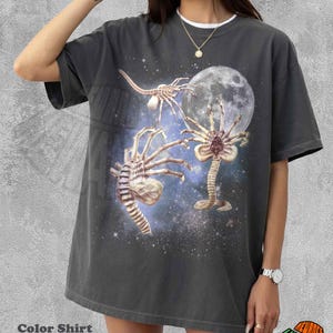 May include: Dark gray t-shirt featuring a graphic of extraterrestrial creatures in a space scene with a full moon. The shirt has a white collar and the text "Color Shirt Pepper".