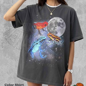 May include: A pepper-colored t-shirt featuring a graphic print of three poison dart frogs in red, blue, and yellow, set against a cosmic background with a large full moon. The shirt has a crew neckline and short sleeves, showcasing a space-themed design.