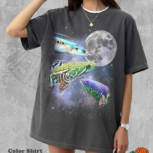 May include: Dark gray t-shirt featuring a vibrant graphic of three colorful fish set against a starry space background with a full moon. The shirt has the text "Color Shirt Pepper" at the bottom.