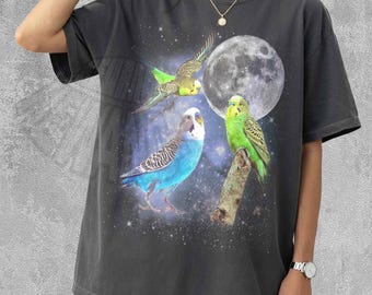 Può includere: Una t-shirt a maniche corte grigio antracite con una stampa grafica di tre pappagallini in un ambiente spaziale stellato con una grande luna piena. Il design include un uccello in volo, uno blu e uno verde appollaiato su un ramo.