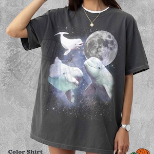 Pode incluir: Uma t-shirt de manga curta cor de pimenta com uma estampa gráfica de três baleias beluga brancas a nadar numa cena espacial estrelada com uma lua grande e detalhada. O design inclui um fundo de nebulosa cósmica com estrelas brancas.