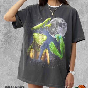 Puede incluir: Camiseta gris oscuro con un gráfico de mantis religiosas, una luna llena y un cielo estrellado. La camiseta tiene el texto "Color Shirt Pepper" en la parte inferior. La camiseta tiene un cuello blanco.