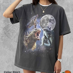 May include: A pepper-colored short-sleeve t-shirt featuring a graphic print of three Godzilla figures against a starry space background with a large full moon. The design includes a roaring Godzilla in the foreground and two others in the distance.