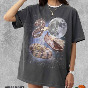 May include: A pepper-coloured short-sleeve t-shirt featuring a graphic print of three snakes and a full moon against a dark, starry space background. The design includes a coiled snake, a snake head, and a winding snake body with detailed scales.