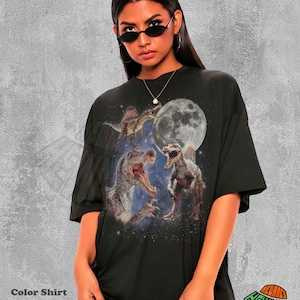 May include: A black short-sleeved t-shirt featuring a graphic print of three dinosaurs in a starry space setting with a large full moon. The design includes a Spinosaurus in the upper left, a large Spinosaurus head, and a standing Spinosaurus.