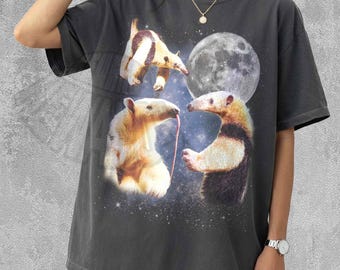 May include: A charcoal grey short-sleeved t-shirt featuring a graphic print of three anteaters against a starry night sky background with a large full moon. The anteaters are depicted in various poses, with one extending a long tongue, set against a cosmic backdrop.