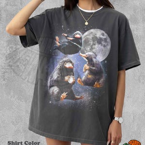 May include: A pepper-colored short-sleeve t-shirt featuring a graphic print of three Niffler creatures from the Fantastic Beasts series. The design includes a large full moon and a starry night sky background. The shirt has a crew neckline.