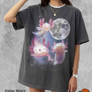 May include: Dark grey t-shirt with a design of three axolotls, a full moon, and a starry night sky. The axolotls are in pink and white. The shirt has a relaxed fit and a white crew neck. The shirt is a pepper color.