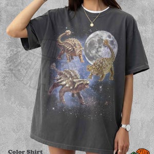 Puede incluir: Camiseta gris oscuro con un gráfico de tres dinosaurios, una luna llena y un cielo estrellado. La camiseta tiene un cuello blanco y el texto "Color Shirt Pepper" en la parte inferior.