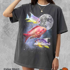 May include: A charcoal gray short-sleeve t-shirt featuring a graphic print of three colorful tropical fish swimming against a dark space background with a large, detailed full moon. The fish are rendered in shades of yellow, pink, and purple.