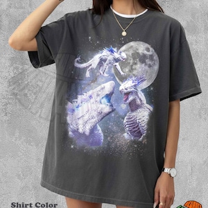 以下が含まれることがあります： ペッパーカラーの半袖Tシャツ。暗い星空の背景に、大きな月と3体の白いトゲのある爬虫類のような生き物が描かれたグラフィックプリントが施されています。デザインには青く光るアクセントと星が含まれています。