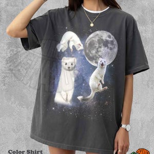 May include: Dark grey t-shirt featuring a whimsical design of white ferrets against a starry space background with a large moon. The shirt has a white collar and the text "Color Shirt Pepper".