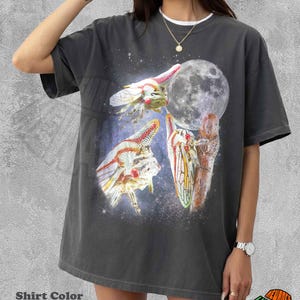 May include: A pepper-colored short-sleeve t-shirt featuring a graphic print of three cicadas against a starry night sky background with a large full moon. The design includes detailed illustrations of the insects with red eyes and translucent wings.