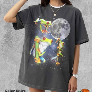 Può includere: T-shirt color pepe con un design stravagante di tre raganelle con occhi rossi su uno sfondo di cielo stellato e luna piena. Le rane sono appollaiate su steli di bambù. La maglietta ha il testo "Color Shirt Pepper".